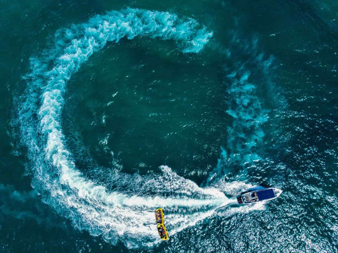 Aerial shot of a speedboat towing a tube, creating a circular wave pattern on the ocean's surface